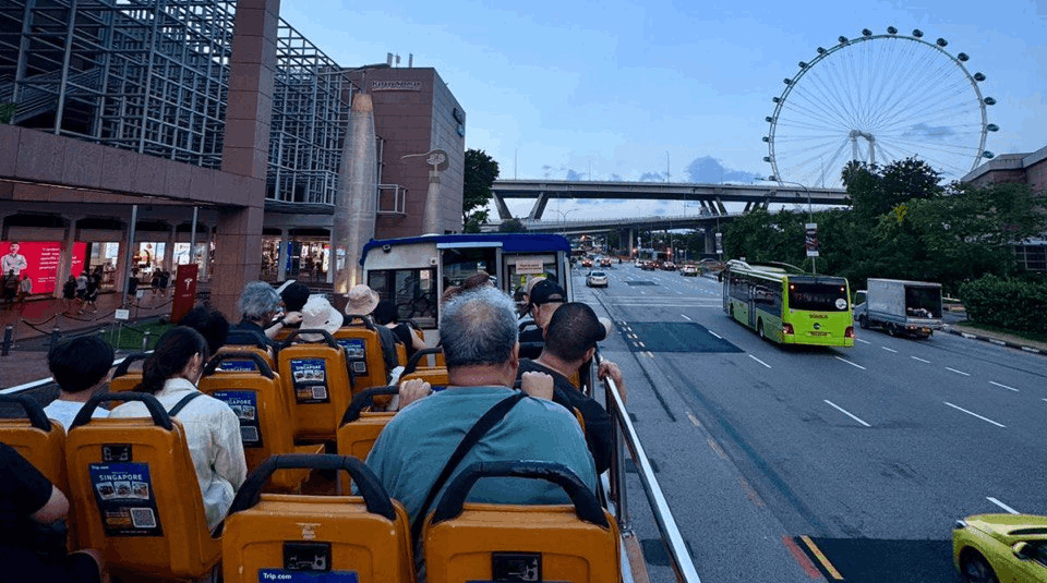 A view from the back of an open-top sightseeing bus in Singapore during the day, showing tourists seated and looking toward the Singapore Flyer and a wide city highway.