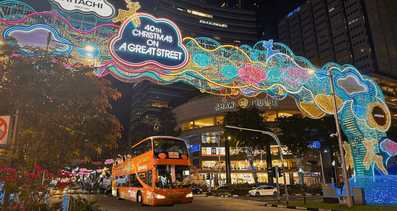 An orange FunVee open-top sightseeing bus passing under the colourful Christmas on A Great Street light arch at Orchard Road, Singapore, during the festive season.