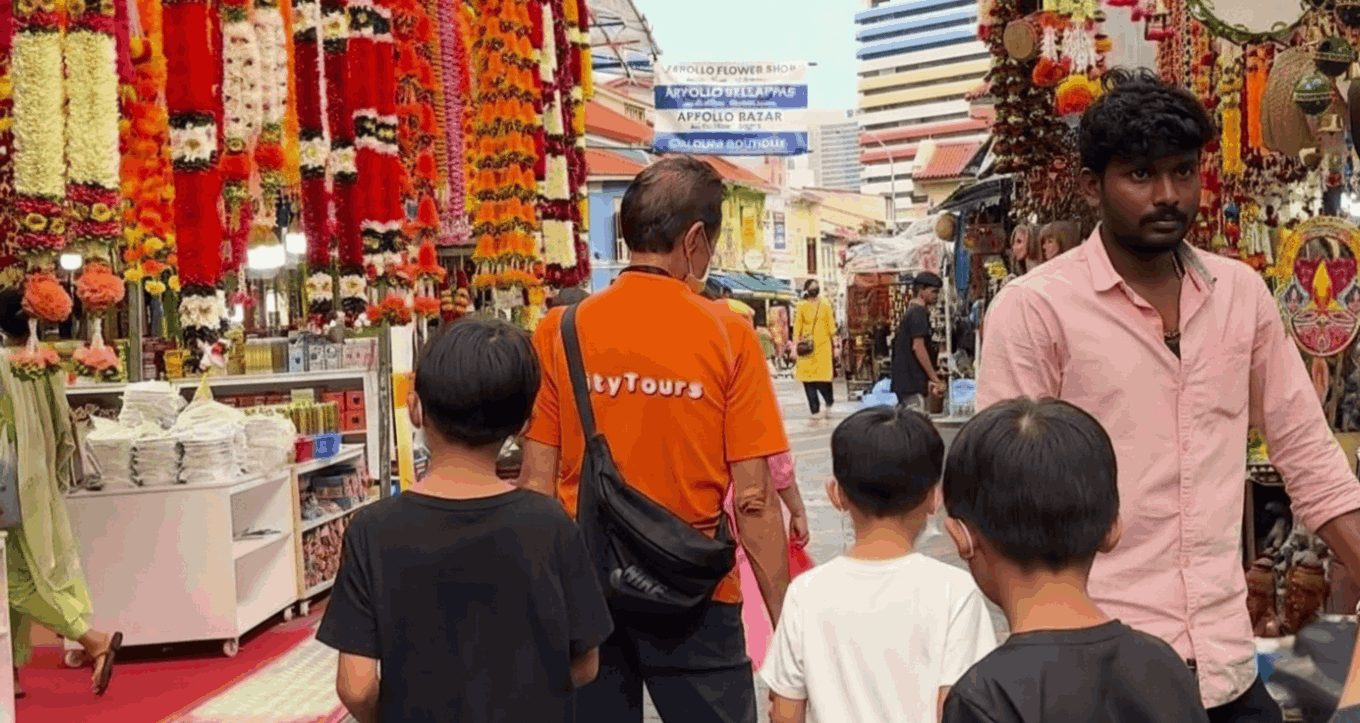 Singapore family walking with City Tourist Guide through the colourful market stalls in Little India selling garlands, spices and traditional goods along a vibrant street.