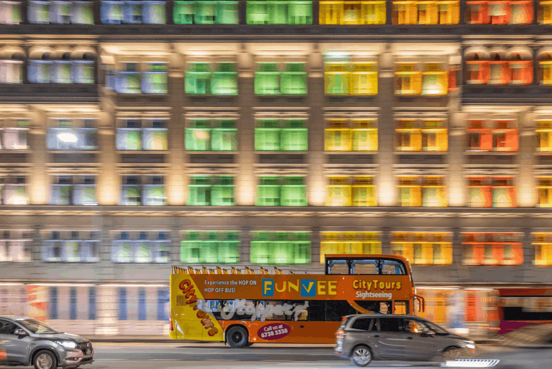 FunVee City Tours open-top hop-on hop-off sightseeing bus driving past the colourful illuminated windows of the Old Hill Street Police Station in Singapore at night.