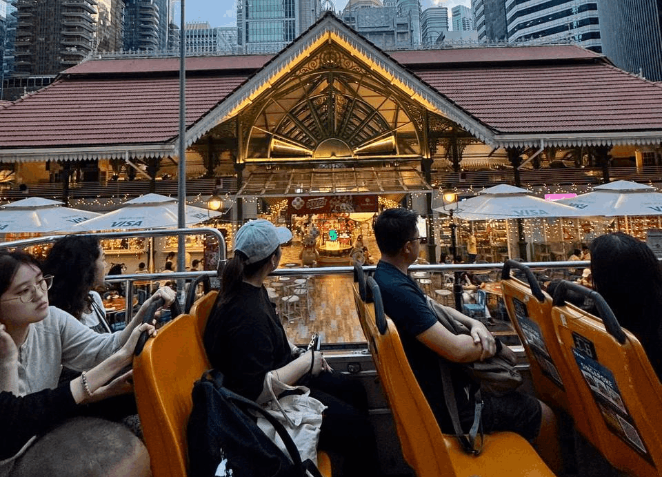 Tourists sightseeing on the upper deck of a FunVee open-top bus passing Lau Pa Sat, with Singapore’s CBD skyline in the background.
