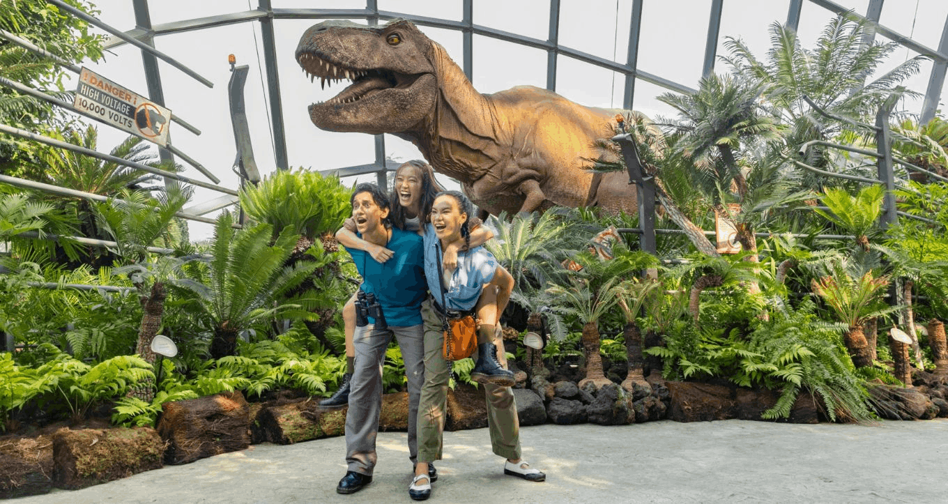 Excited people, smiling and looking thrilled, in front of a large, realistic Tyrannosaurus Rex dinosaur model amidst lush tropical plants at the Jurassic World Experience in Gardens by the Bay.