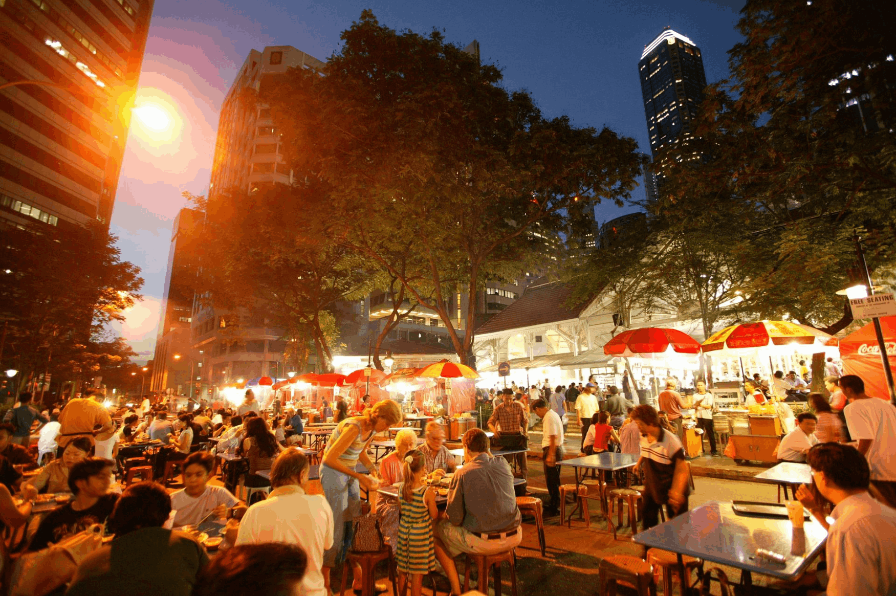 Evening scene at Lau Pa Sat in Singapore with people dining outdoors along Satay Street, surrounded by smoky barbecue stalls and city skyscrapers.