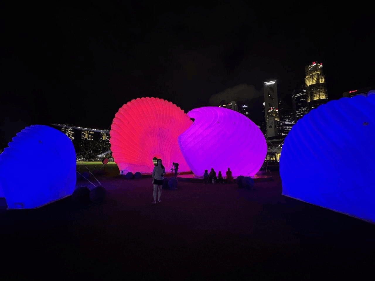 Large, illuminated pink and blue shell-shaped inflatable art installations at night, set against the backdrop of the Singapore Central Business District skyline.