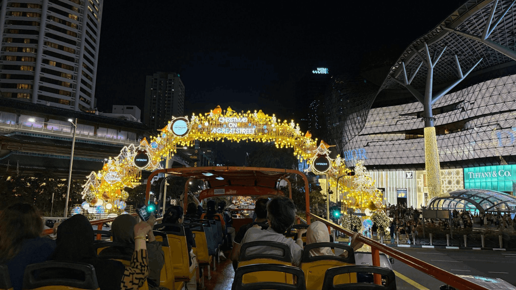 Passengers on an open-top bus tour approaching the main "Christmas on a Great Street" arch in front of ION Orchard at night.