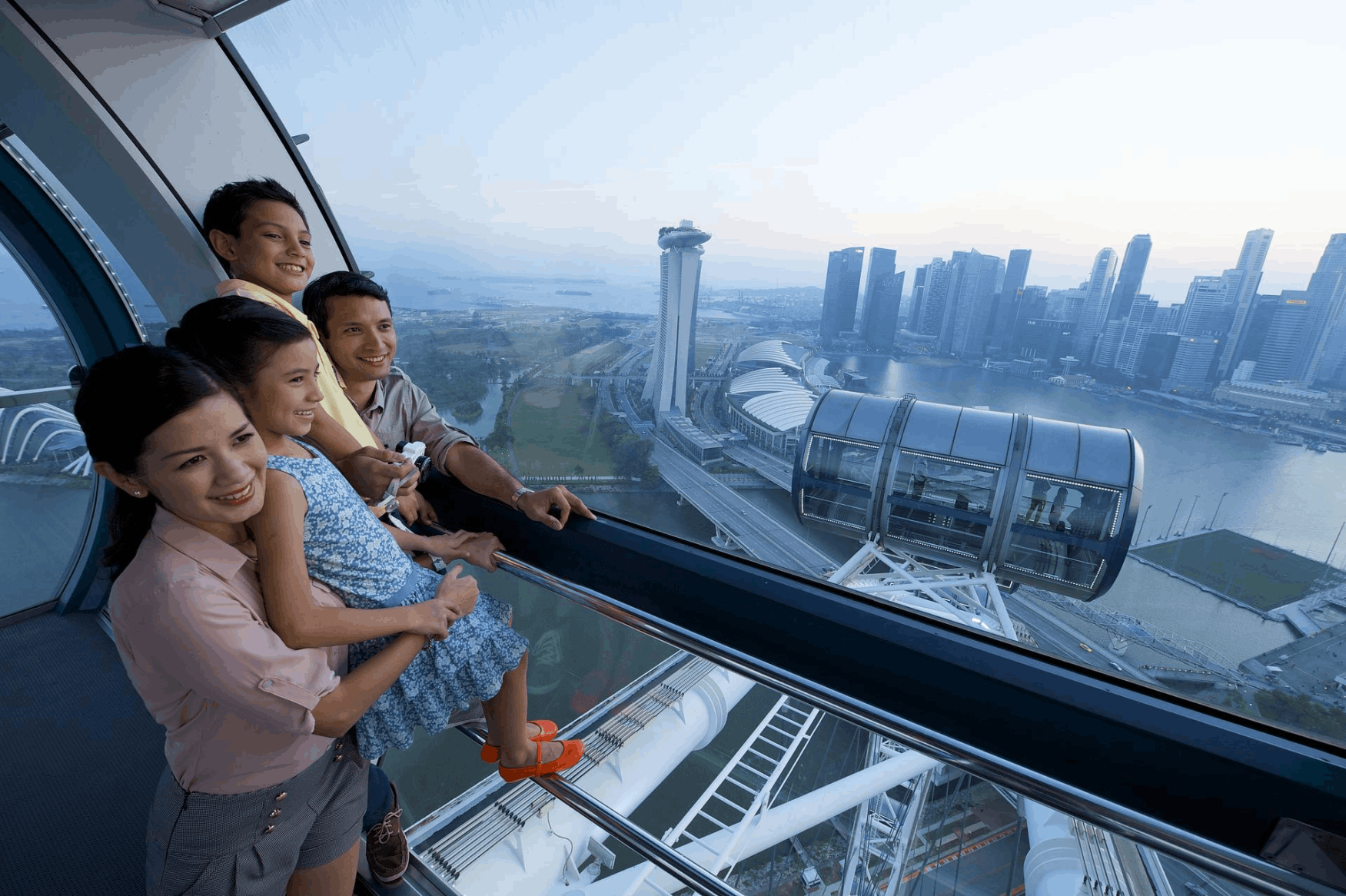 A happy family, including two children, looking out from a capsule of the Singapore Flyer, enjoying the panoramic view of the Singapore city skyline and waterfront. themed buildings, palm trees, and colourful storefronts under a large transparent canopy.