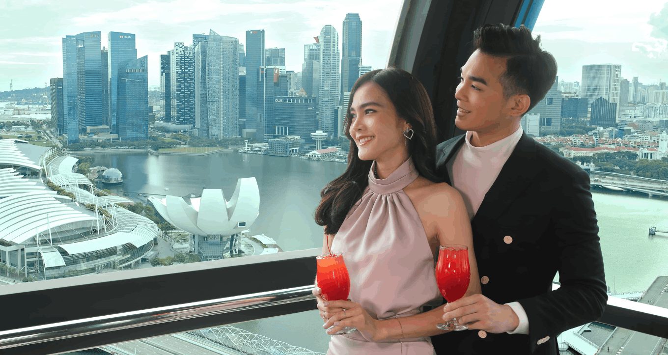 A stylish couple smiling and holding Singapore Sling cocktails inside a Singapore Flyer capsule, enjoying the panoramic view of the Marina Bay skyline.