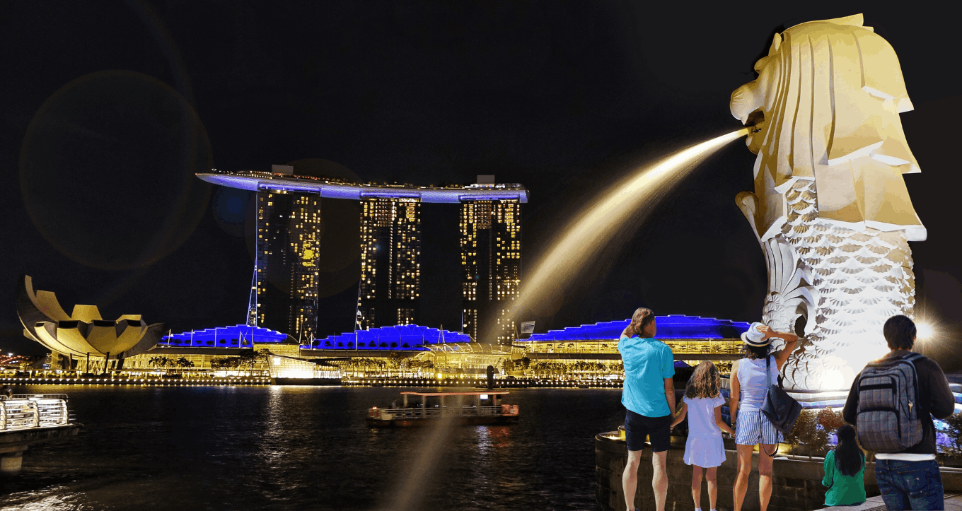 Visitors walking along Merlion Park at night with the illuminated Merlion statue and Singapore’s city skyline glowing in the background.