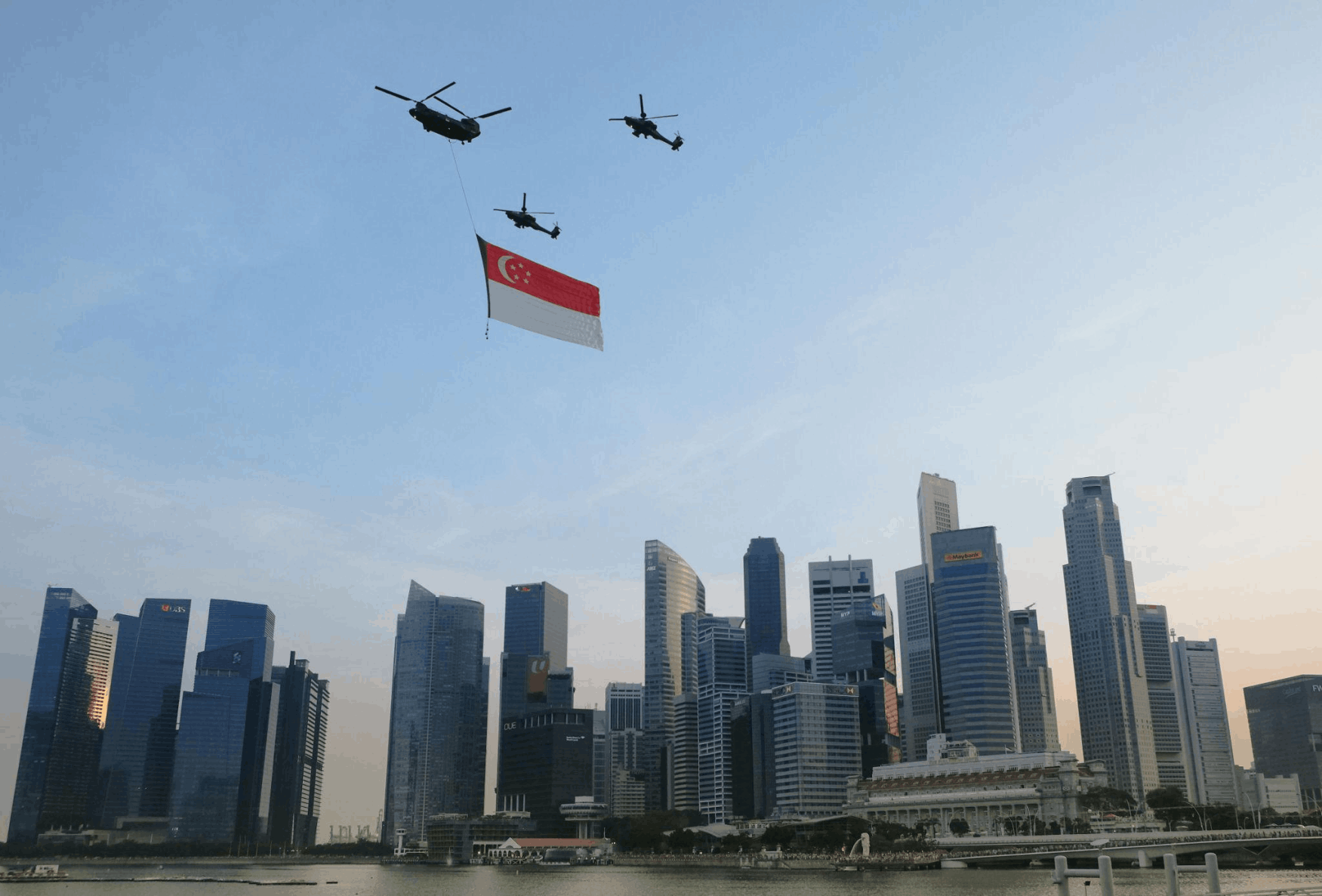 Helicopters flying over Marina Bay with a large Singapore flag during National Day celebrations, with city skyscrapers in the background