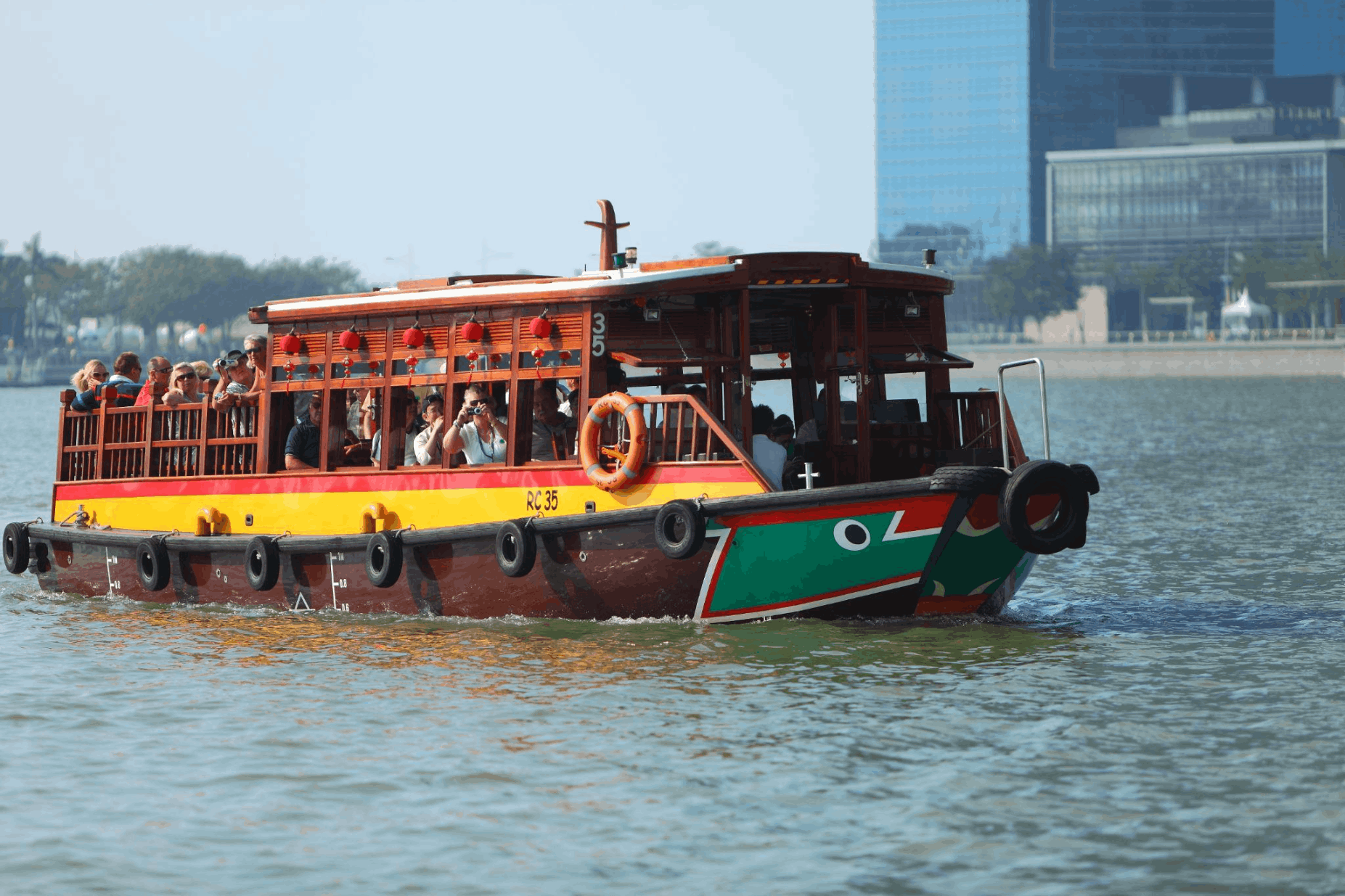 A Singapore River Cruise bumboat, painted green, yellow, and red with decorative red lanterns, filled with tourists taking pictures while sailing past the city skyline.