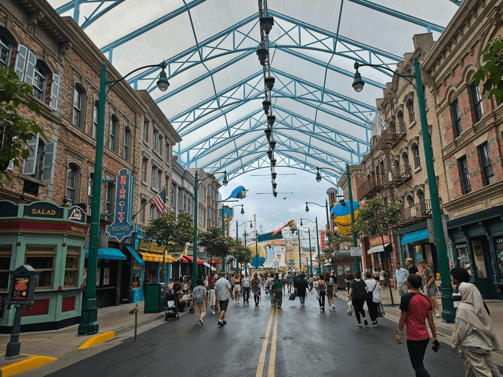 Visitors walking along Hollywood Street at Universal Studios Singapore, surrounded by themed buildings, palm trees, and colourful storefronts under a large transparent canopy.